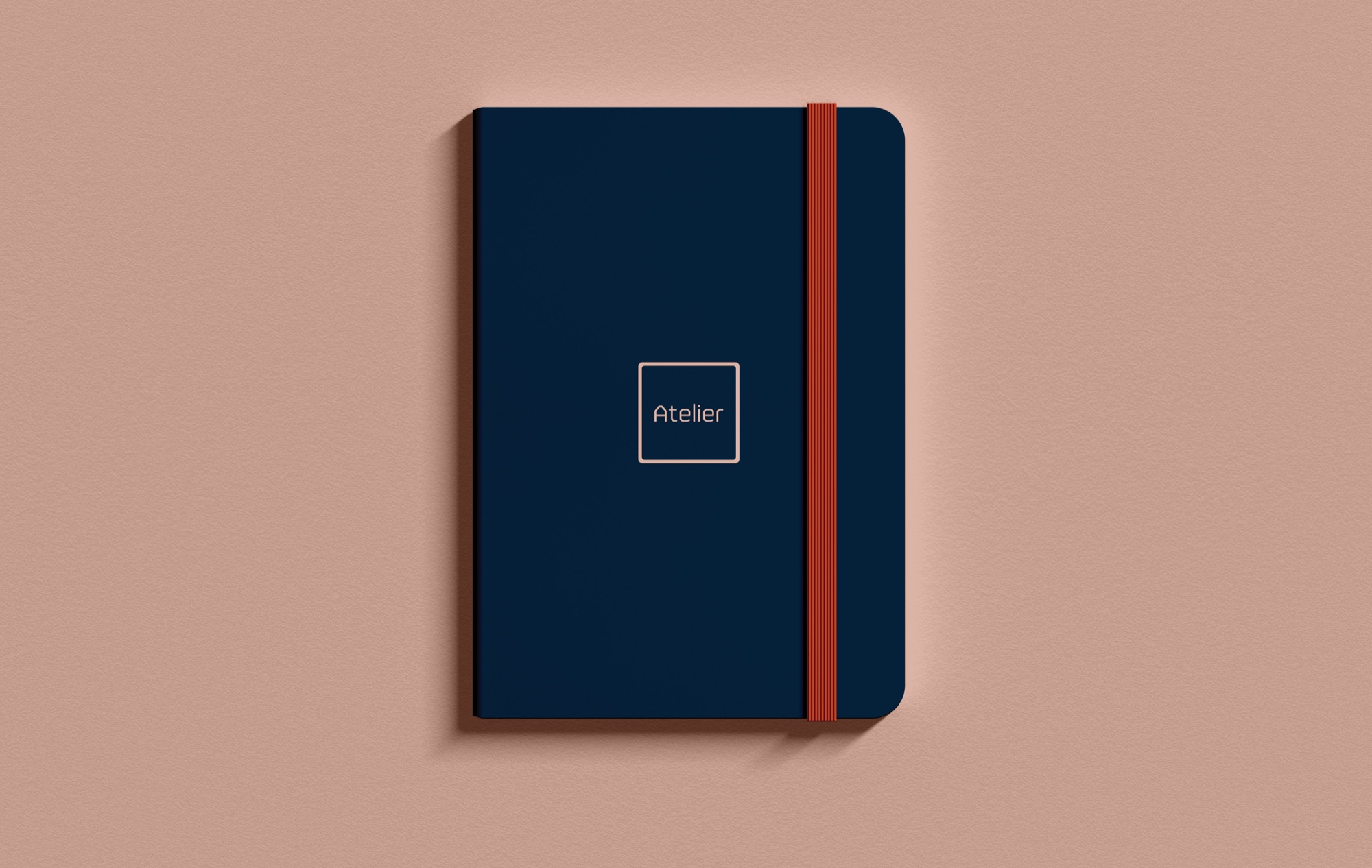 Navy hardback notebook with the blush Atelier square logo centred and a ribbed orange elastic closure, photographed flat on a blush ground