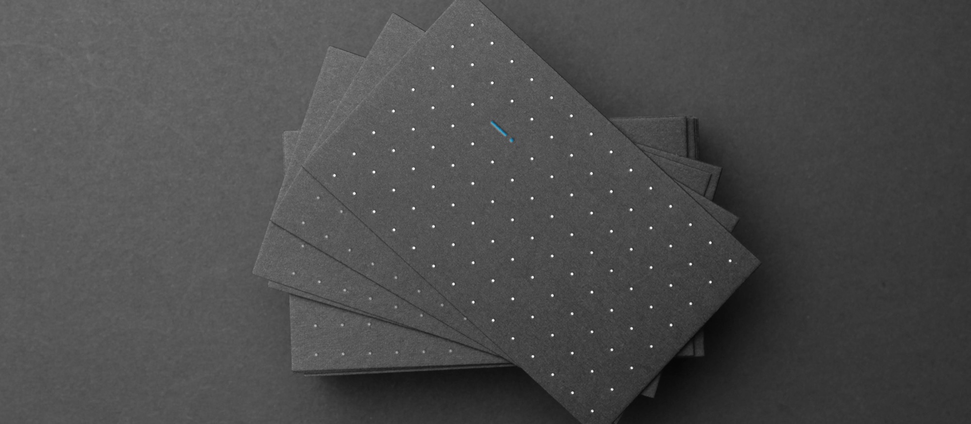 A fan of charcoal business cards on a dark grey ground, each printed with a white dot grid and a single cyan mark standing out among the dots