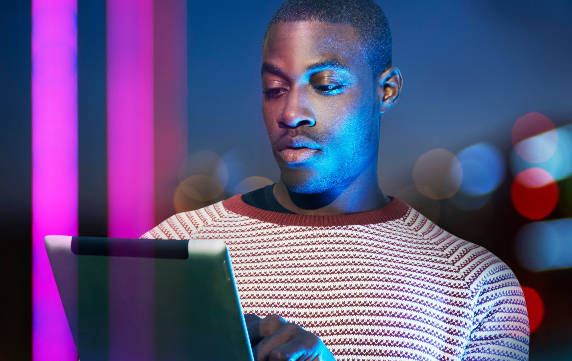 A man in a patterned jumper reading a tablet, lit blue and magenta by neon tubes and city bokeh