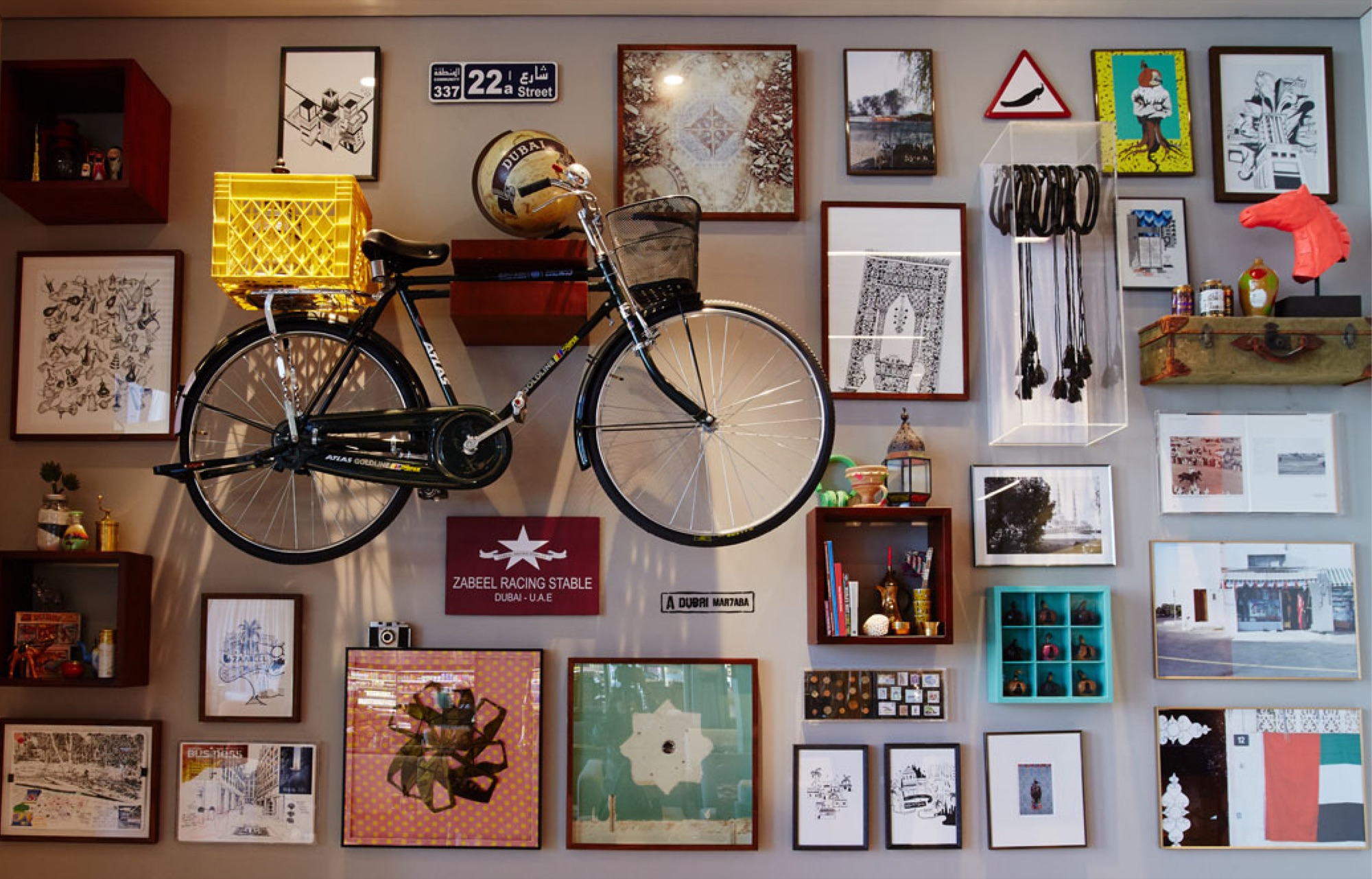 Gallery wall: a black Atlas bicycle mounted centrally with a yellow crate on its rack, surrounded by framed drawings and photographs of Dubai, a 22a Street sign, a Zabeel Racing Stable plaque, open display boxes of local objects, a vintage suitcase and a coral horse-head sculpture