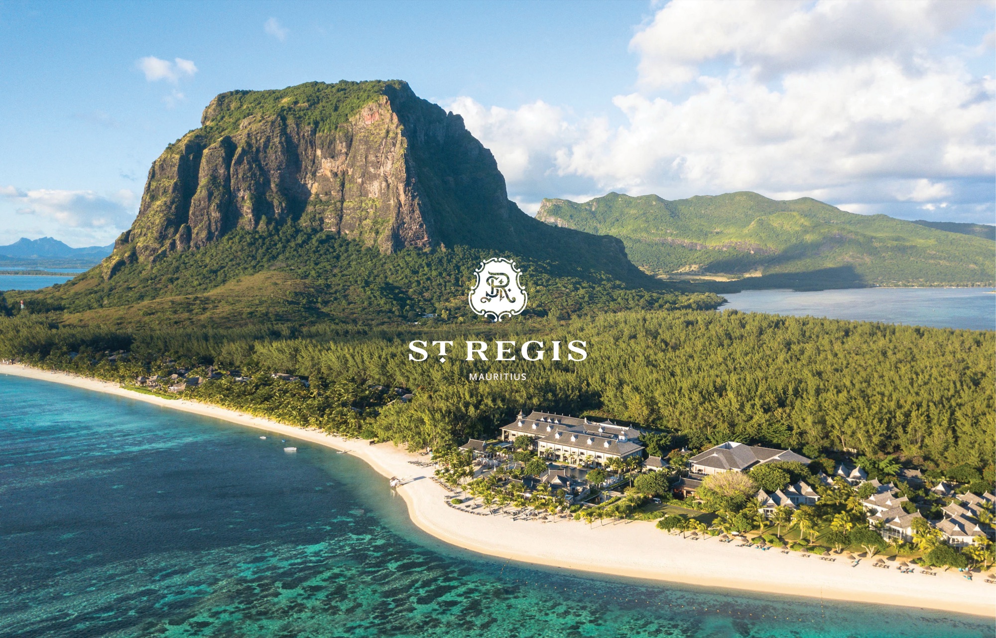 Aerial view of Le Morne: the UNESCO-listed basalt mountain rising over forest, a white sand spit and turquoise reef, with the resort's colonial-style manor houses along the shore and the St Regis Mauritius crest and wordmark set over the scene