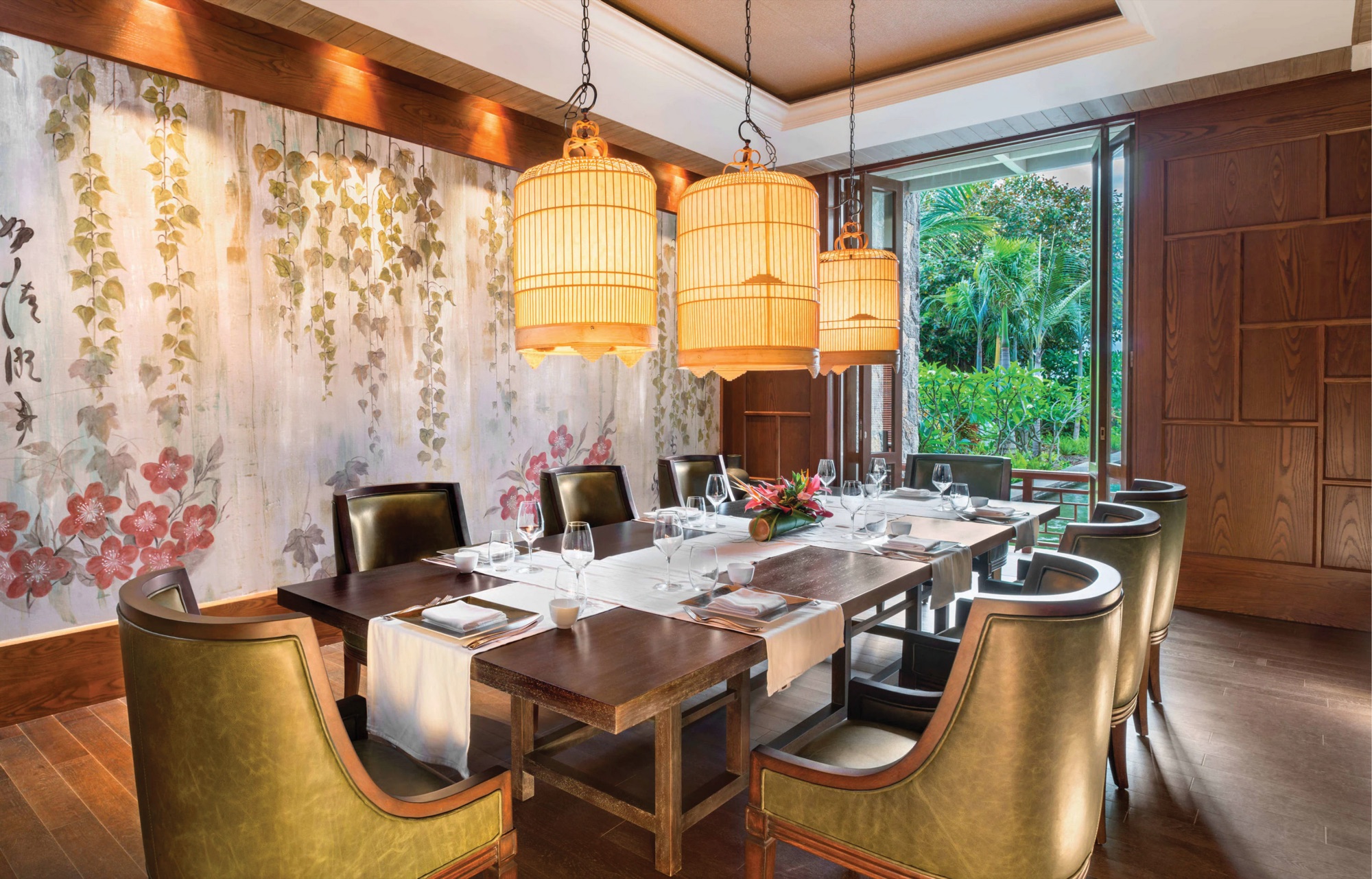 Private dining room with three woven birdcage pendants over a long timber table, the wall painted with trailing vines and red blossom, and folding doors open to the garden