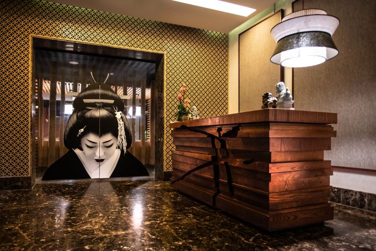 The Atsuko restaurant entrance: a geisha painted across mirrored doors framed in brass, a gold lattice screen wall, a stepped timber host desk bound in black cord, and a drum pendant over polished marble