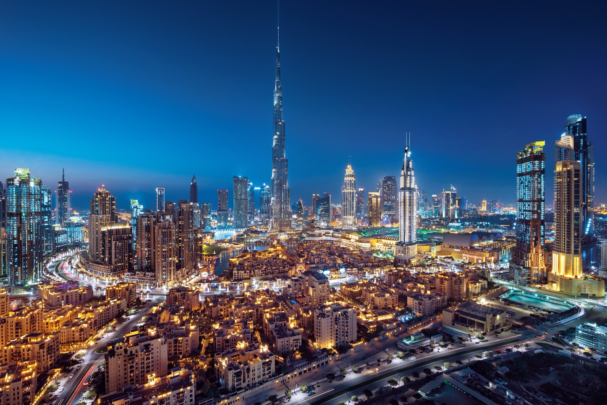 Downtown Dubai at dusk: the Burj Khalifa lit against a deep blue sky, the Address hotel towers and Dubai Mall below, and light trails running along Sheikh Zayed Road
