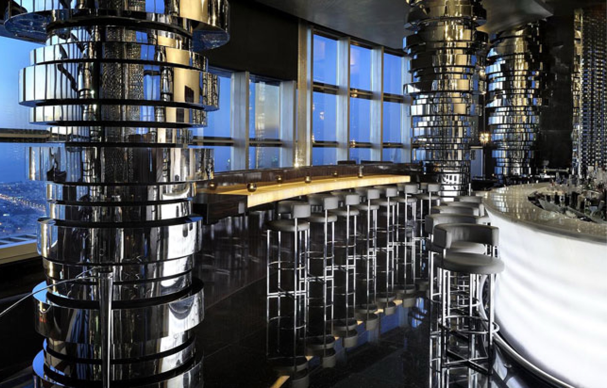 NEOS bar on the upper floor: tiered polished steel and brass columns flanking a curved onyx-lit counter with chrome stools, reflected in a black mirror floor, city lights through full-height glazing at dusk