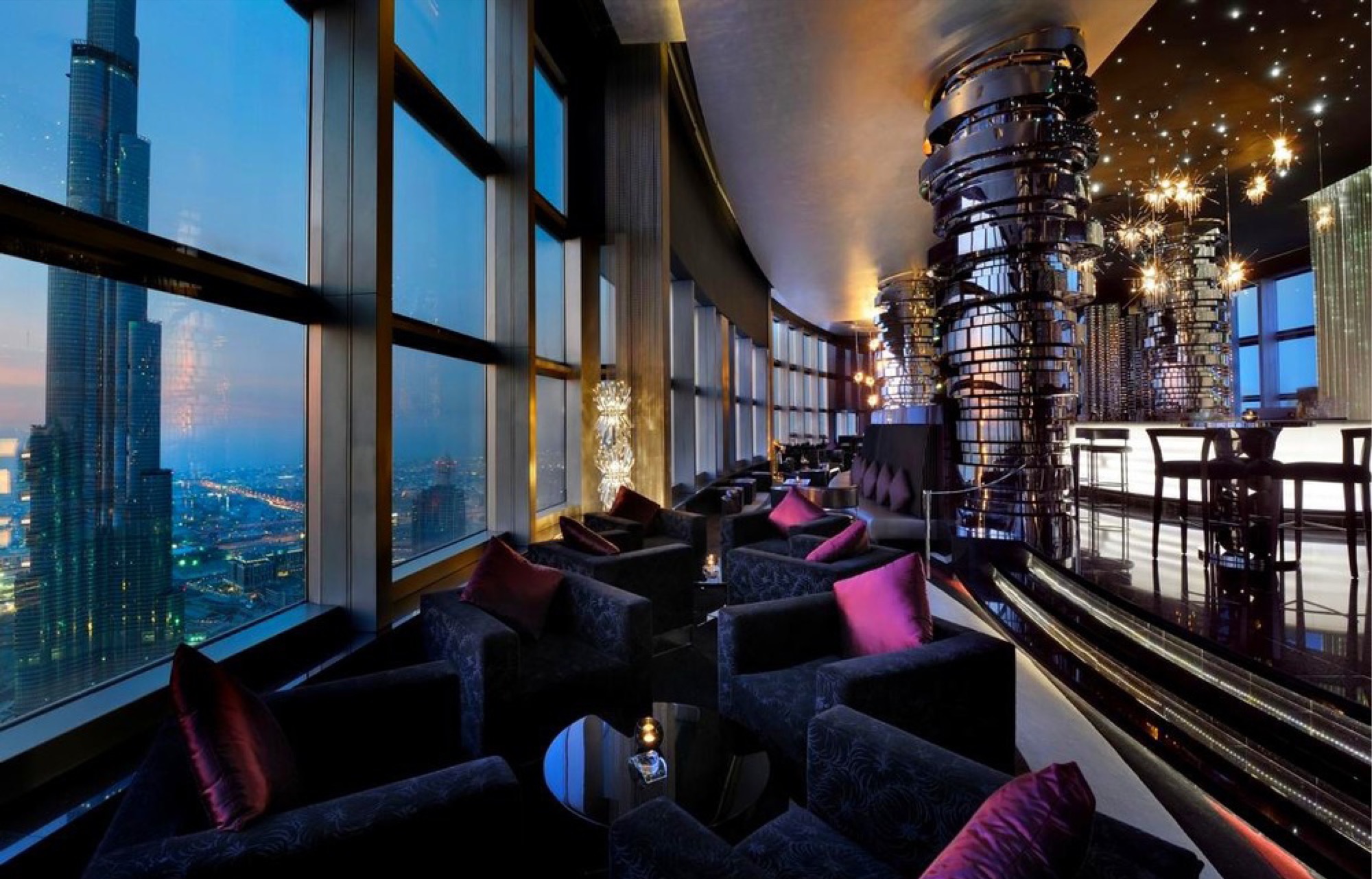 NEOS lounge at dusk: velvet armchairs in charcoal with magenta and burgundy cushions along a curved wall of glazing looking onto the Burj Khalifa and the city lights, mirrored steel columns and starburst chandeliers under a dark ceiling of fibre-optic points